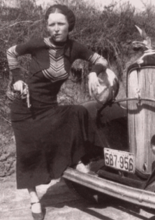 Bonnie Parker steht an ein Auto gelehnt, eine Zigarre im Mund und eine Pistole in der Hand